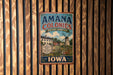 a vintage-style metal sign with the text "AMANA COLONIES HERITAGE LANDSCAPE" and "IOWA" printed on it, hanging on a wooden wall.