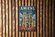 a vintage metal sign with the text "Amiens Cathedral" and "France" written on it, hanging on a wooden wall.