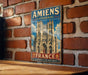 A vintage metal sign featuring an illustration of the Amiens Cathedral in France, mounted on a brick wall.