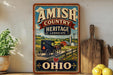 a vintage-style sign for Amish Country Heritage Landscape in Ohio, with a farm scene and a horse-drawn carriage.