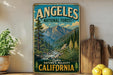A vintage-style metal sign for the Angeles National Forest in California, featuring a scenic mountain landscape with pine trees, a river, and a waterfall.