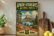 a vintage-style poster of the Apache-SITGREaves National Forests in Arizona, with a scenic mountain landscape and a lake in the background. The poster is displayed on a wall, surrounded by various kitchen items such as a cutting board, a knife, a spoon, and a bowl.