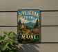 A vintage metal sign for the Appalachian Trail in Maine, featuring a scenic mountain landscape with pine trees and a trail sign.