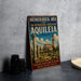 A vintage-style metal sign on a table, featuring an illustration of an ancient building and the text "ARCHAEOLOGICAL AREA AND THE PATRIARCHAL BASILICA OF AQUILEIA ITALY DISCOVER 2000 YEARS OF HISTORY WHERE THE PAST LIVES ON".