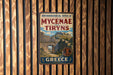 a vintage-style metal sign with the text "ARCHAEOLOGICAL SITES OF MYCENAE AND TIREYS GREECE" displayed on a wooden wall.