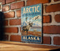 A vintage-style sign for the Arctic National Wildlife Refuge in Alaska, featuring an illustration of a moose and a bird in a mountainous landscape.