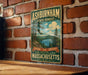 A vintage-style metal sign for Ashburnham State Forest, featuring a scenic landscape with pine trees and a lake, mounted on a brick wall.