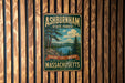 A vintage metal sign for Ashburnham State Forest, featuring a scenic landscape with pine trees, a lake, and mountains.