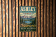 A vintage metal sign for the Ashley National Forest in Utah, featuring a scenic mountain landscape with pine trees and a lake.