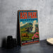 A vintage-style metal sign for Aso Park, Japan, featuring a cow grazing in a mountainous landscape with a volcano in the background.