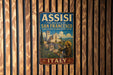 A vintage metal sign with the text "ASSISI THE BASILICA OF SAN FRANCISCO AND OTHER FRANCICAN SITES" and an illustration of a person in a robe and hat, standing in front of a church.