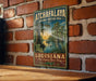 A vintage-style metal sign for the Atchafalaya National Heritage Area in Louisiana, featuring a scenic illustration of a bayou landscape with a bird and a palm tree.
