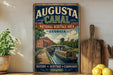 a vintage-style metal sign for the Augusta Canal National Heritage Area in Georgia, with a painting of a canal and buildings in the background.