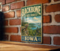 A vintage metal sign for the Backbone State Forest in Iowa, featuring a scenic landscape with a lake and mountains.