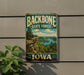 A vintage-style metal sign for the Backbone State Forest in Iowa, featuring a scenic landscape with a lake and mountains.