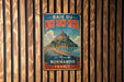 A vintage metal sign with the text "BAIE DU MONT SAINT-MICHEL" and "La Merveilleuse de Normandie FRANCE" is displayed on a wooden wall.