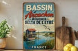 A vintage-style metal sign advertising the Bassin d'Arçon, a coastal town in France, with a scenic image of a small boat on the water and a picturesque landscape in the background.