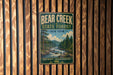 A vintage metal sign for Bear Creek State Forest in New York, featuring a scenic illustration of a forest landscape with a river and waterfall.