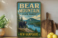 A vintage-style metal sign for Bear Mountain State Forest in New Hampshire, featuring a black bear standing on a rocky outcropping overlooking a lake surrounded by pine trees.