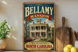 A vintage-style metal sign for Bellamy Mansion in Winston-Salem, North Carolina, featuring an illustration of the mansion surrounded by trees and flowers.