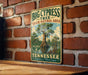 A vintage-style sign for the Big Cypress Tree State Natural Area in Tennessee, featuring a large cypress tree and a river.