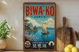 a vintage-style sign for Bwa-ko, Japan, with a scenic illustration of a boat sailing on a lake and a temple in the background. The sign also includes text that reads "Bwa-ko Japan" and "Japan's largest lake", indicating the location and its significance.