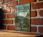 a vintage-style metal sign with a scenic forest landscape, mounted on a brick wall.