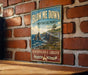 A vintage-style metal sign with the text "BLOW ME DOWN PROVINCIAL PARK NEWFOUNDLAND & LABRADOR ADVENTURE AWAITS" is displayed on a brick wall.