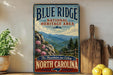 A vintage-style sign for the Blue Ridge National Heritage Area in North Carolina, featuring a scenic mountain landscape with pine trees and pink flowers.