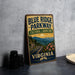 A vintage-style sign for Blue Ridge Parkway, featuring a scenic mountain landscape with a winding road and flowers, is displayed on a gray countertop next to some black camera lenses.