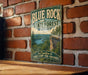 A vintage-style metal sign for the Blue Rock State Forest in Ohio, featuring a scenic landscape with a cliff and a river.