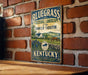 A vintage-style metal sign with the text "Bluegrass Cultural Landscape of the Inner South" and "Kentucky" printed on it, hanging on a brick wall.