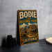A vintage-style sign for Bodie State Historic Park in California, featuring an illustration of a small town or village.