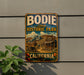 A vintage-style metal sign for Bodie State Historic Park in California, featuring an illustration of a small town and a sign that reads "California".