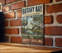 A vintage-style metal sign for Botany Bay Plantation Heritage Preserve, featuring a scenic illustration of a coastal landscape with palm trees and a bird.