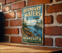A vintage-style metal sign with a canoe and a scenic landscape of a lake and mountains, mounted on a brick wall.