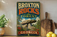 A vintage-style sign for the Broxton Rocks Forest in Georgia, featuring a scenic landscape with a large rock formation and surrounded by trees and flowers.