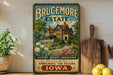 a vintage-style sign for "Brucemore Estate" with a large illustration of a house surrounded by flowers and greenery. The sign is displayed on a shelf next to a wooden cutting board and some potted plants.