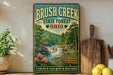 A vintage-style sign for Brush Creek State Forest in Ohio, featuring a scenic illustration of a waterfall surrounded by lush greenery and flowers.