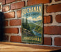 A vintage-style metal sign for the Bucanan State Forest in Pennsylvania, featuring a scenic landscape with pine trees, a river, and mountains.