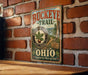 A vintage-style metal sign for the Buckeye Trail in Ohio, featuring a brick wall backdrop and a scenic illustration of the trail.