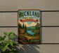 A vintage metal sign for Buckland State Forest in Massachusetts, featuring a scenic landscape with pine trees, a lake, and mountains.