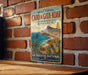 A vintage-style metal sign with a beach scene and mountains in the background, mounted on a brick wall.