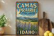 a vintage-style sign for Camas Prairie, a cultural landscape in Idaho, with a scenic mountain landscape and purple wildflowers in the foreground.