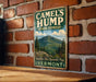 A vintage metal sign for Camel's Hump State Forest in Vermont, featuring a mountain landscape with pine trees and a cloudy sky.