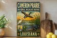 a vintage-style sign for the Cameron Prairie National Wildlife Refuge in Louisiana, depicting various wildlife species such as ducks, all set against a scenic landscape.