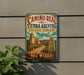 A vintage-style metal sign with a colorful illustration of a desert landscape, featuring a building and a cross, mounted on a wall.