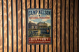 A vintage metal sign for the Camp Nelson National Monument in Kentucky, featuring an illustration of a log cabin and a wagon, mounted on a wooden wall.
