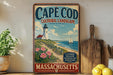 A vintage-style metal sign for Cape Cod, Massachusetts, featuring a lighthouse, flowers, and a sailboat on the water.