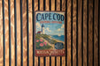 A vintage metal sign for Cape Cod, Massachusetts, featuring a lighthouse and a beach scene.